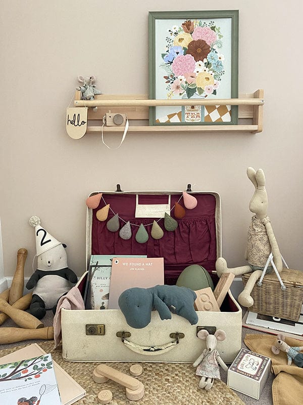 A vintage suitcase open, against a wall with toys and books spilling out of it, Maileg mice, a Maileg bunny, a soft panda with a party hat on, below a spice rack shelf with a maileg mouse, a hello wooden pennant, a wooden toy camera and our Boho Floral vase art print in a green frame.