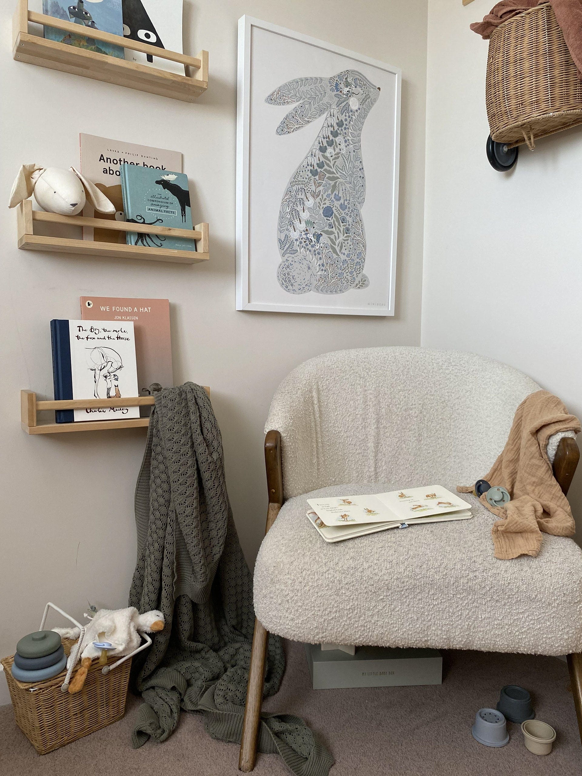 Reading nook in a bedroom or nursery in neutral colours, with books in spice rack shelves, some toys and a green blanket on the floor. An Olli Ella Luggy hanging on the wall above the cosy of white seat with darker wooden legs, with the Floral Bunny Art print above it.
