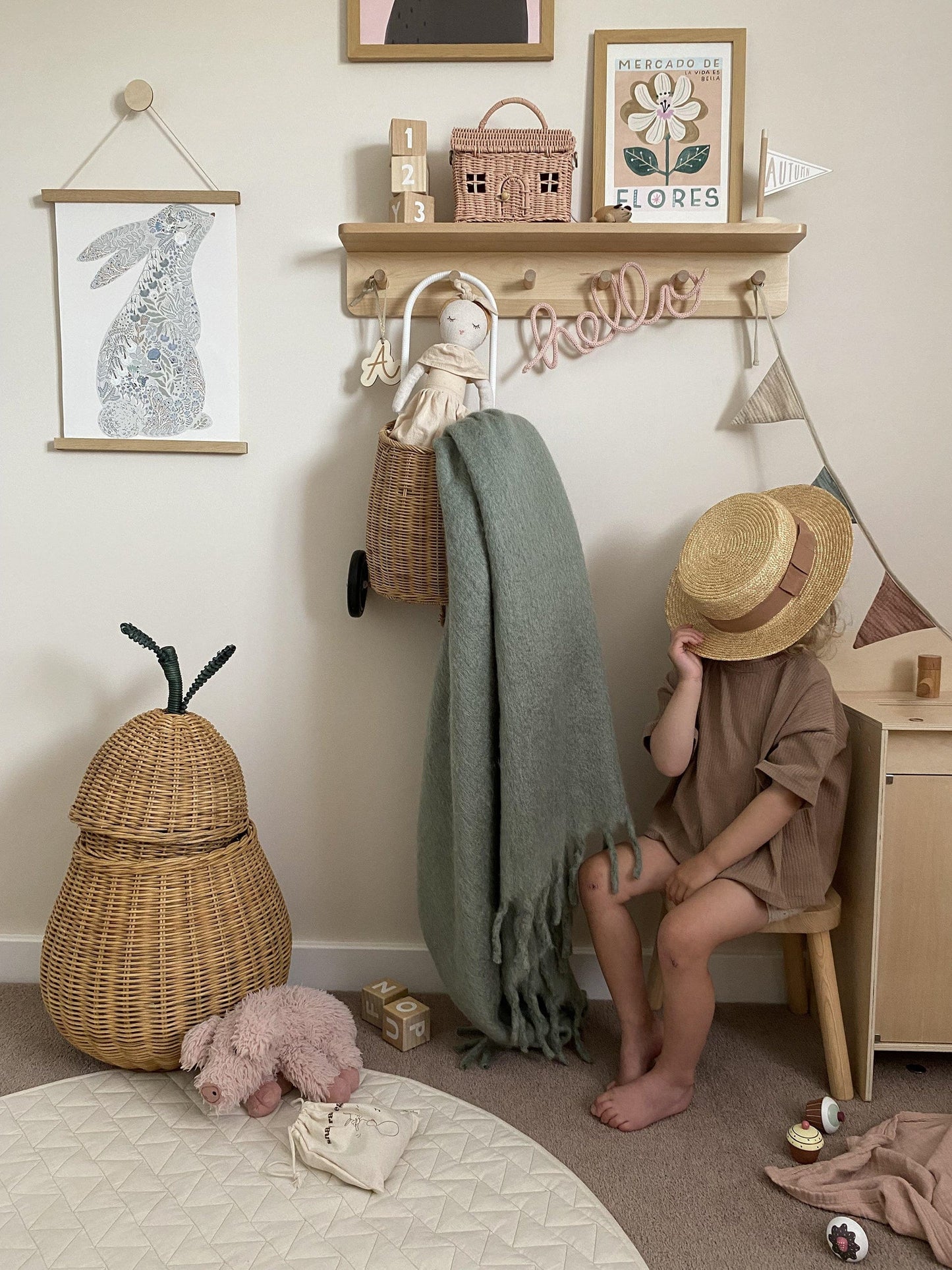 A child sitting on a wooden stool hiding their face with a straw hat, next to a ferm living pear basket and below a wooden shelf with hooks. An Olli Ella luggy hangs from the hooks with a doll and blanket hanging out of it, a wire-wrapped 'hello' sign, and bunting. On the shelf are wooden blocks, an Olli Ella Casa Clutch, a wooden flag saying autumn, and our Mercado de Flores art print in a frame. Hanging from an oak hanger from a round decorative hook is our floral bunny art print in blue.