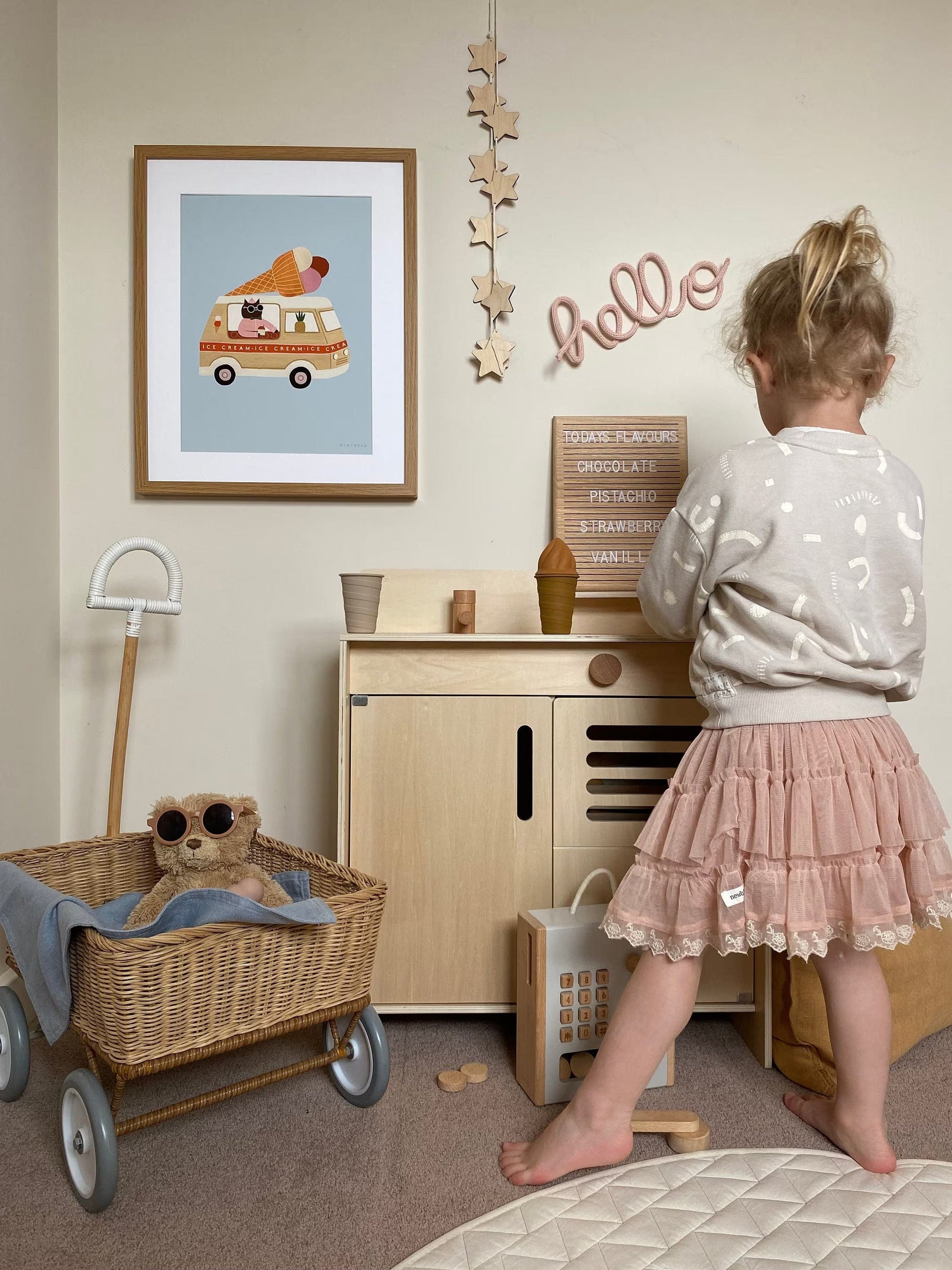 Girl in a pink skirt and beige sweater playing at a Liewood Mario Wooden toy kitchen, with liewood ice cream toys. With an Olli ella wagon with a teddy sitting in the bottom left. On the wall above the kitchen is our ice cream van art print, mounted in a wooden frame, some hanging wooden stars and a wrapped wire hello sign.