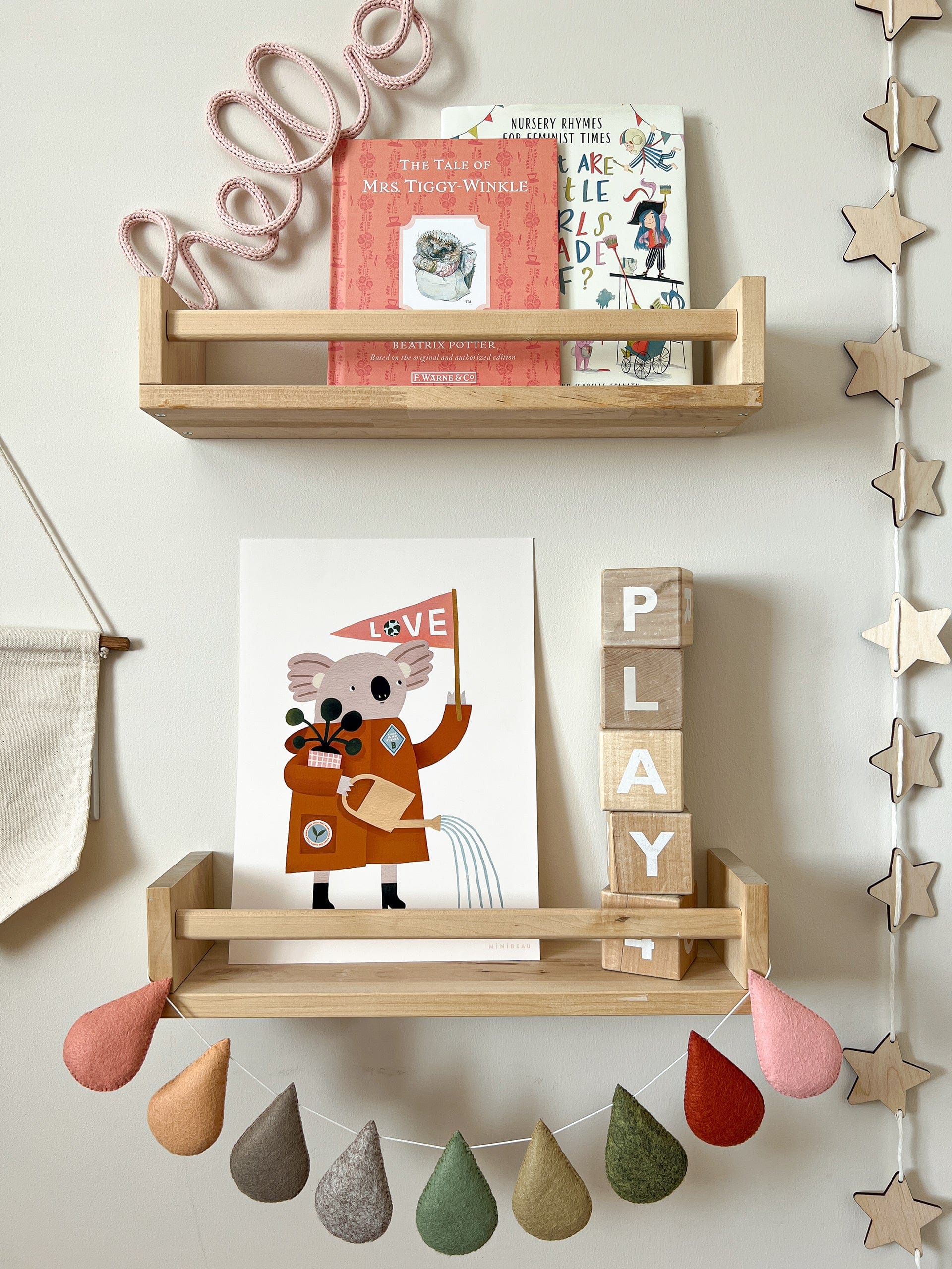 2 spice rack style shelves one above the other with a string of wooden stars to the right. On the top shelf art 2 books and a wrapped wire word HELLO. On the lower shelf art wooden blocks stacked to spell play and our Love our planet koala art print, with raindrop bunting in autumnal colours hanging on the front.