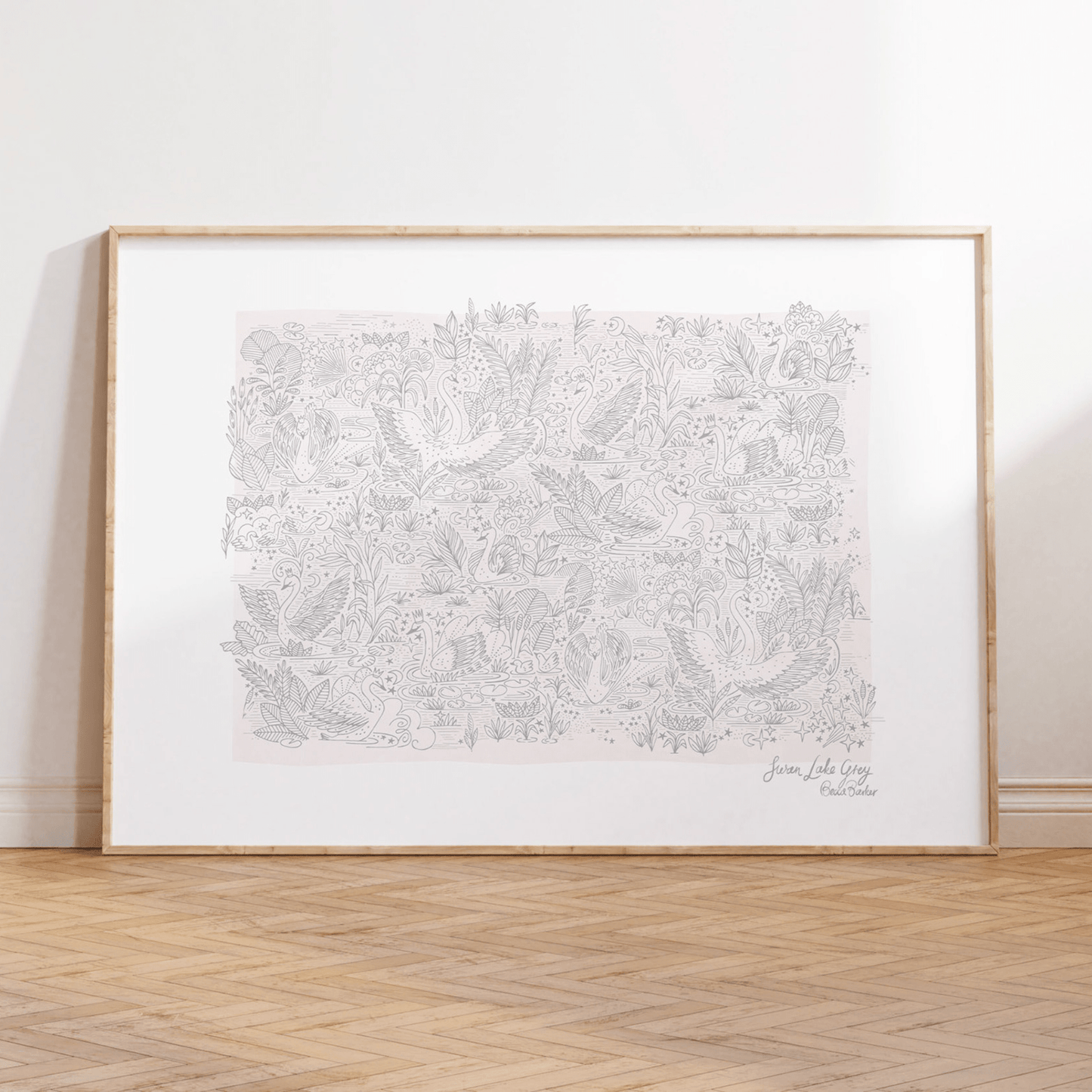 Art print in alight wood frame leaning against a neutral wall, standing on parquet flooring. Our swan lake grey art print features, fine grey lined drawing on a pale pink background. Showing detailed foliage, and elegant swans in small crowns gliding over a star filled lake.