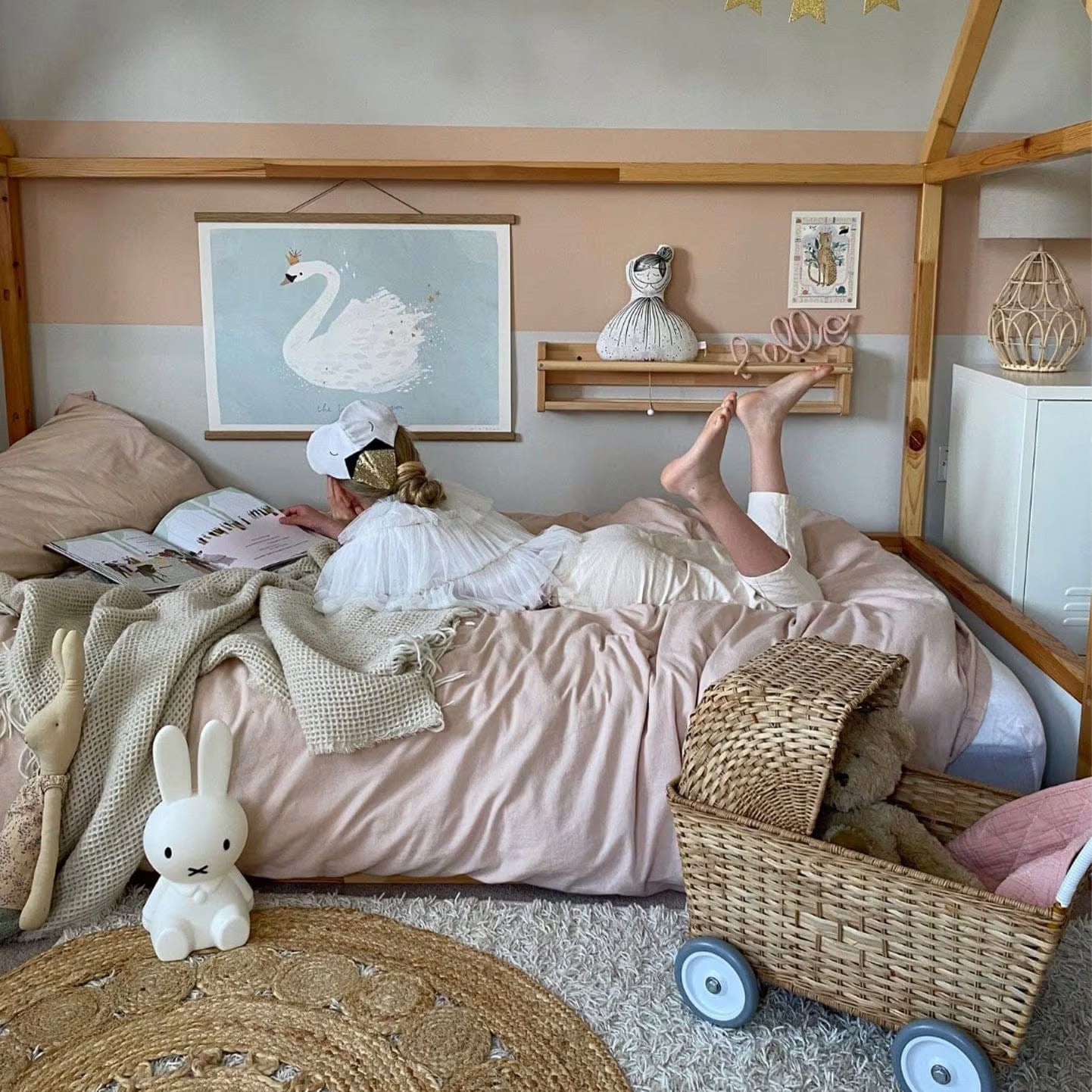 Looking in on a girl laying on her house frame bed reading. On the floor is a Miffy lamp and an Olli Ella Strolley. The wall is decorated with a wide horizontal pink stripe. Above the girl on the wall is our The Wonder of you art print in an oak hanger. Above a spice rack shelf which has a wrapped wire hello sign on is our wild, wonder world leopard print.