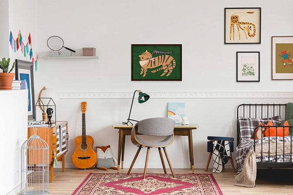 An older childs room, with a black metal frame bed, with a traditional red rug on the floor and a wooden desk with grey chair. On the wall is a selection of art prints in black frames of varying sizes, our Forever Fearless art print in green, Giraffe art print, wood art print and it's how I roll art print.