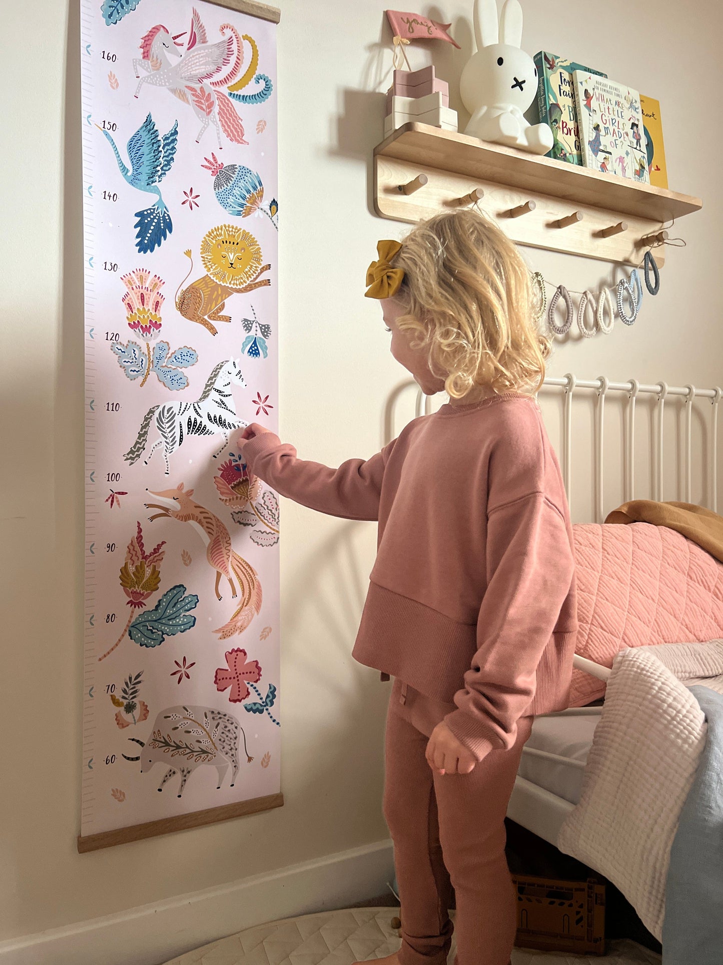 Blonde girl pointing at the zebra on the folk carnival height chart. Vintage metal framed bed in the background and a wooden shelf with peg hooks with a miffy lamp on it and raindrop bunting