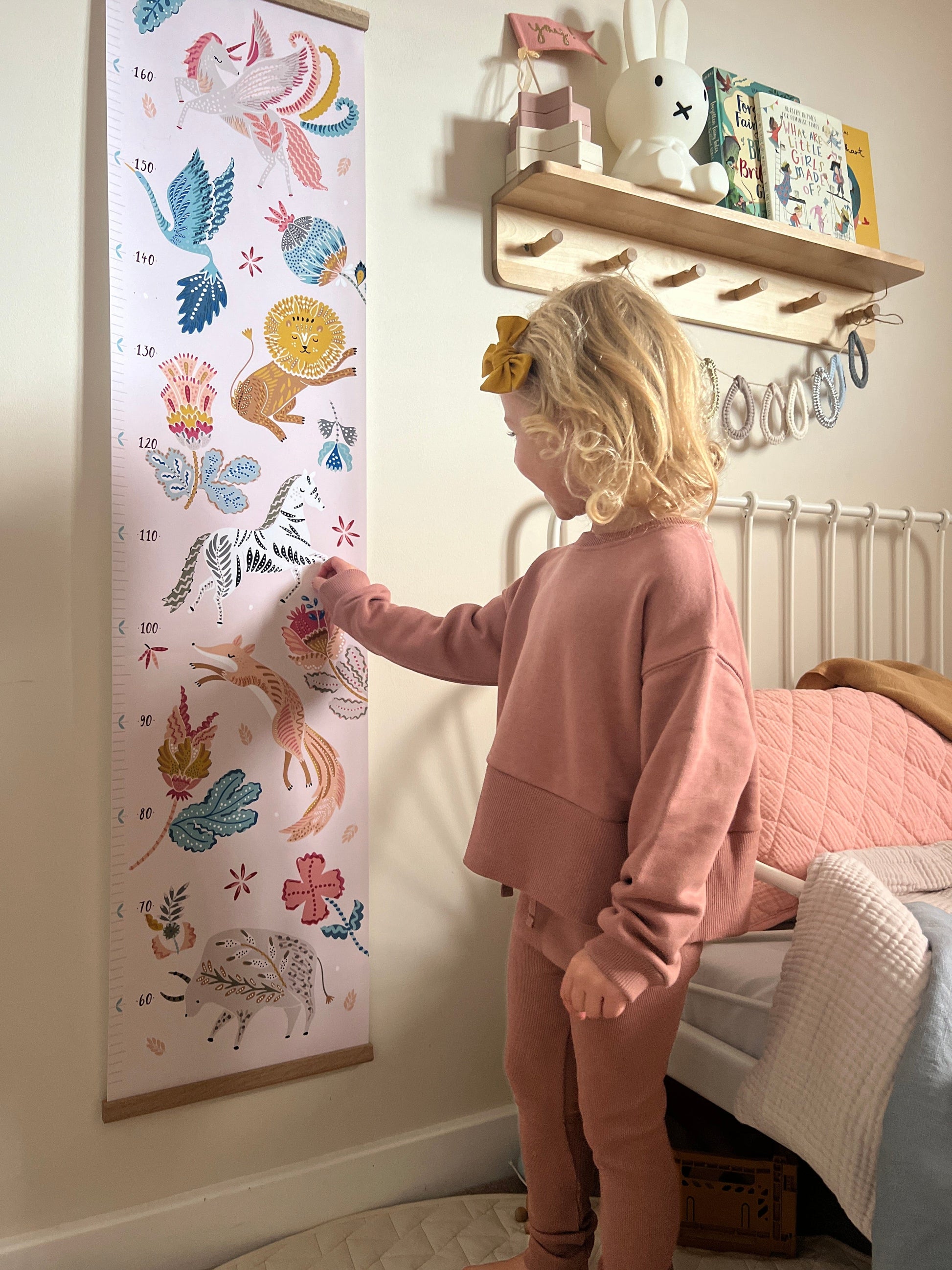 Blonde girl pointing at the zebra on the folk carnival height chart. Vintage metal framed bed in the background and a wooden shelf with peg hooks with a miffy lamp on it and raindrop bunting