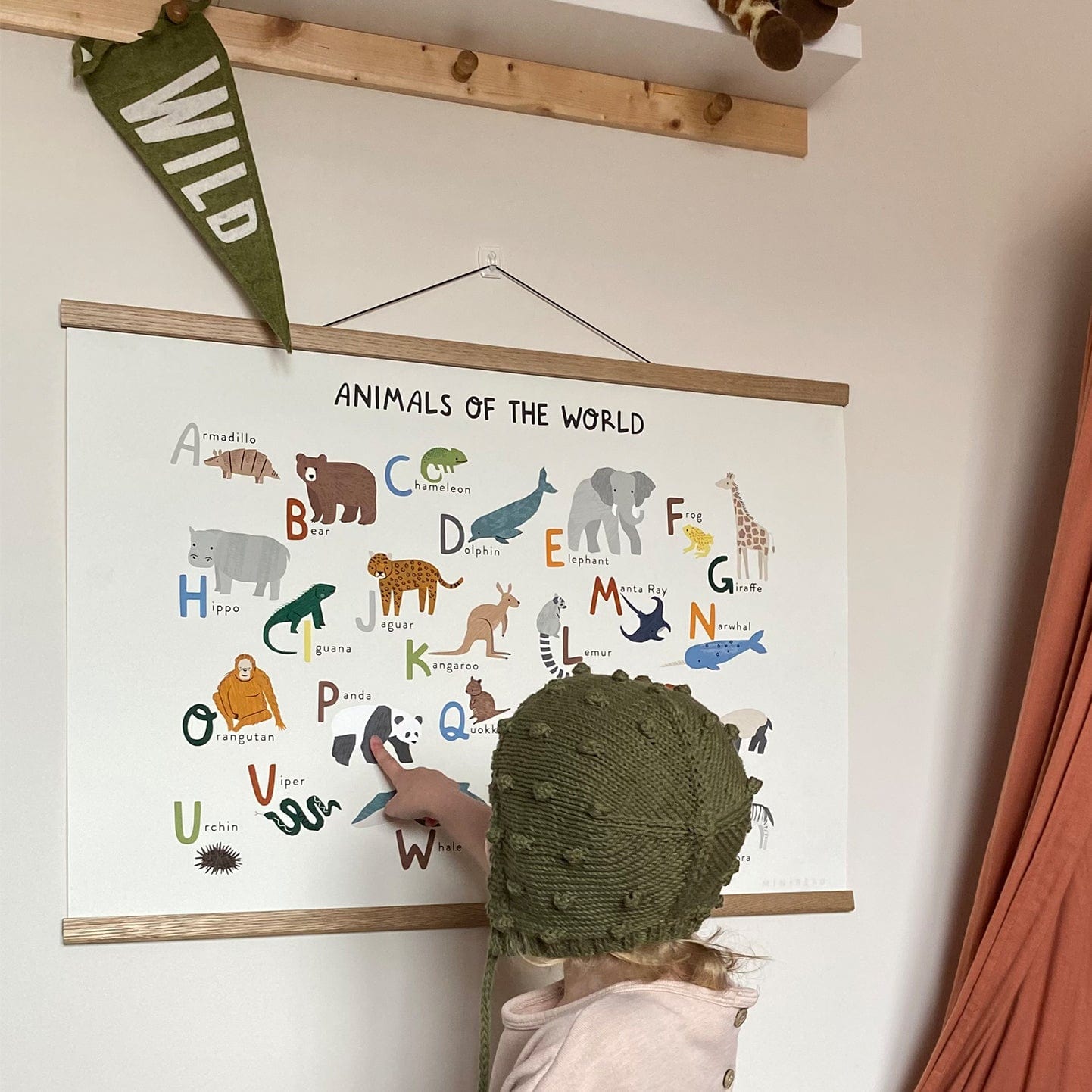 A photo of a girl in a green woollen bonnet pointing at a panda on the animals of the world art print, which is hanging in an oak hanger, with a small triangle flag with the word wild on it. The art print consists of pictures of animals from around the world representing each letter of the alphabet. Has text at the top saying animals of the world in large font and the text - An alphabet of just 26 of the wondrously, amazing 8.7 million animal species living on earth in brackets underneath.