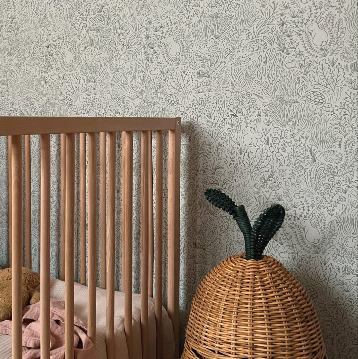 Corner of a natural wooden cot with peach bedding next to a ferm living pear basket in front of a wall with garden doodles sage wallpaper.