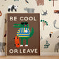 Art print in a light wood frame on a parquet floor leaning against a wall with our paper cut animals wallpaper hung on it. Our be cool or leave art print show a crocodile in a yellow and blue striped jumper and sunglasses, standing on his hind legs holding a skateboard, with the words be cool or leave in off-white on a brown background.