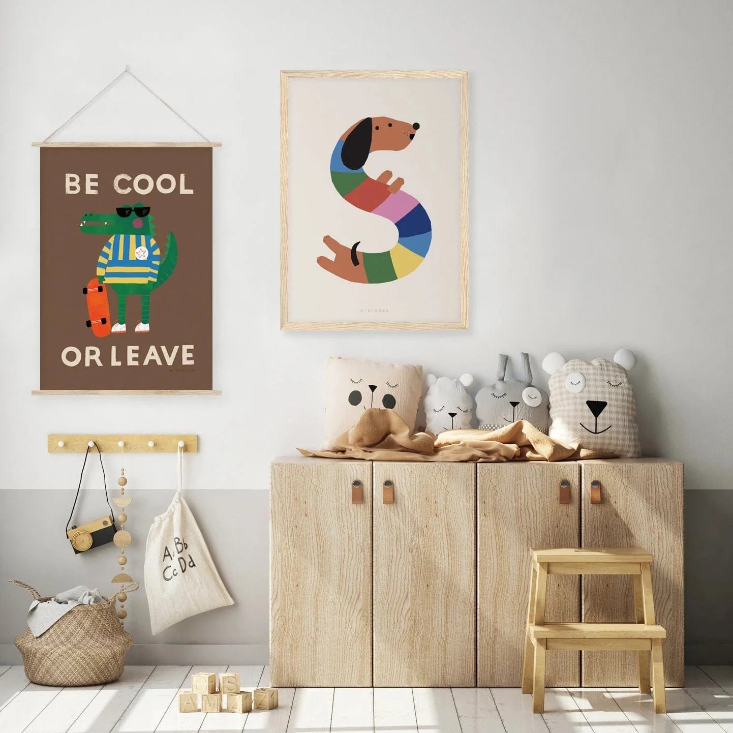 A playroom with low level cupboards with blankets and animal cushions on top , woth wooden toys hanging from a hook on the wall  next to the cupboard. Hung on the wall are two prints be cool and leave in an oak hanger, and Happy Alphabet S in a light wood frame. 