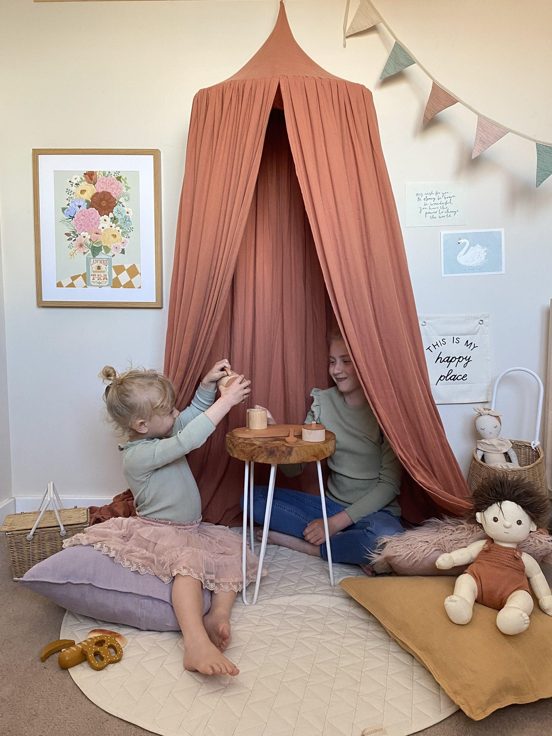 Two girls sitting on floor cushions under a rust coloured canopy, having a tea party with a wooden tea set from liewood and an Olli Ella doll in the foreground with the Boho Floral Vase art print, the wonder of you art print, and  my wish for you art print on the wall behind.