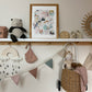 A wooden and white painted shelf in a kids space with hooks on the underside. With bunting, a rainbow mobile, a pink bonnet and a Olli Ella luggy with a doll, all in muted tones, underneath. On the shelf sit an Olli Ella Casa Clutch, a soft panda, a framed Crayon Wilderness art print, booties and a rattan framed mirror.