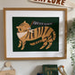 Art print in an oak frame above a shelf that has an I am an explorer wooden pennant, blue kids binoculars, and a wooden catapult on it, and a cream triangle flag with the word explore in brown above the print. Our forever fearless art print features a smiling tiger wearing a white bandana with orange polka dots. It's head is turned over it's shoulder and it is holding a black triangle flag with the words FOREVER FEARLESS in white, it it's mouth. All on a green background.