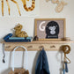 A wooden shelf with wooden pegs underneath with a Liewood blue dinosaur towel, and liewood blue cat skipping rope and Olli Ella Luggy with wooden alphabet blocks in hanging, with books, liewood mustard bre sandals, a toy gorilla, a wooden yoyo and our G is for gorilla art print in an wooden frame. On the wall above is our Giraffe art print and a wooden cutout of the word PLAY.