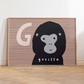 Art print in a light wood frame, leaning against a white wall, standing on parquet flooring. Our G is for Gorilla art print features a smiling cartoon gorilla from the shoulders up, on a brown wood effect background with a large G in a pale brown next to the Gorilla's head.