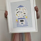 Child in a muted rainbow coloured skirt, holding up a mounted art print in a white frame. Our Good Vibes Gumball Art Print shows a vintage style gumball machine with balloons on either side on a blue background with white line check design. Inside the glass ball of the gumball machine are the words good vibes, sunglasses, roller skates, a cat in sunglasses, ice cream, a rainbow and more.
