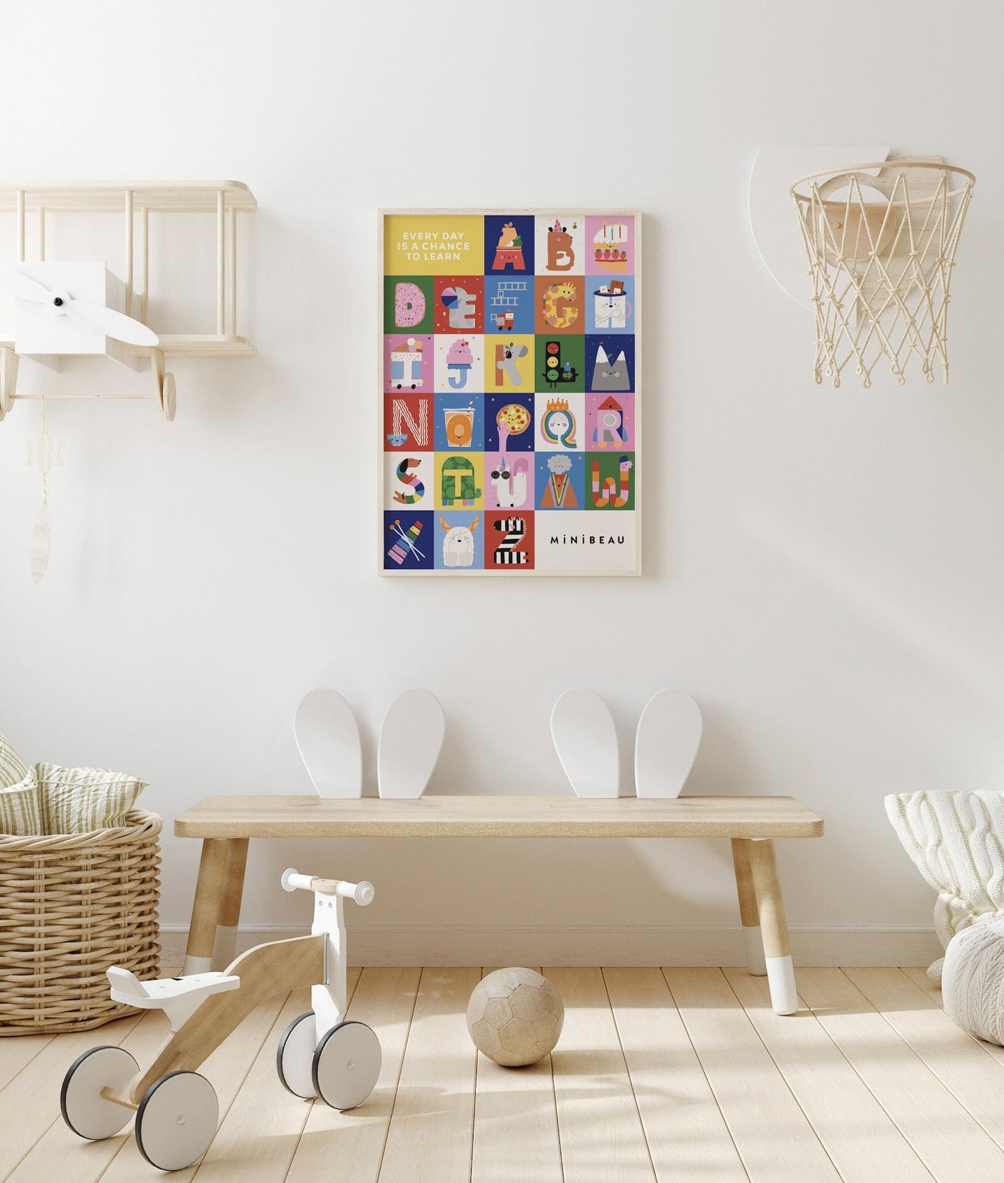 A neutral childs room with a wooden aeroplane shelf and white basketball hoop on the wall, with a wooden trike and a football on the floor in the foreground. Above a wooden bench with 2 sets of bunny ears as seat backs, is our happy alphabet art print in a light wood frame. Our Happy Alphabet Art Print shows all of our Happy Alphabet letters in alphabet form, with EVERY DAY IS A CHANCE TO LEARN in white in a yellow square in the top left, and Minibeau in black in a white square in the bottom right.