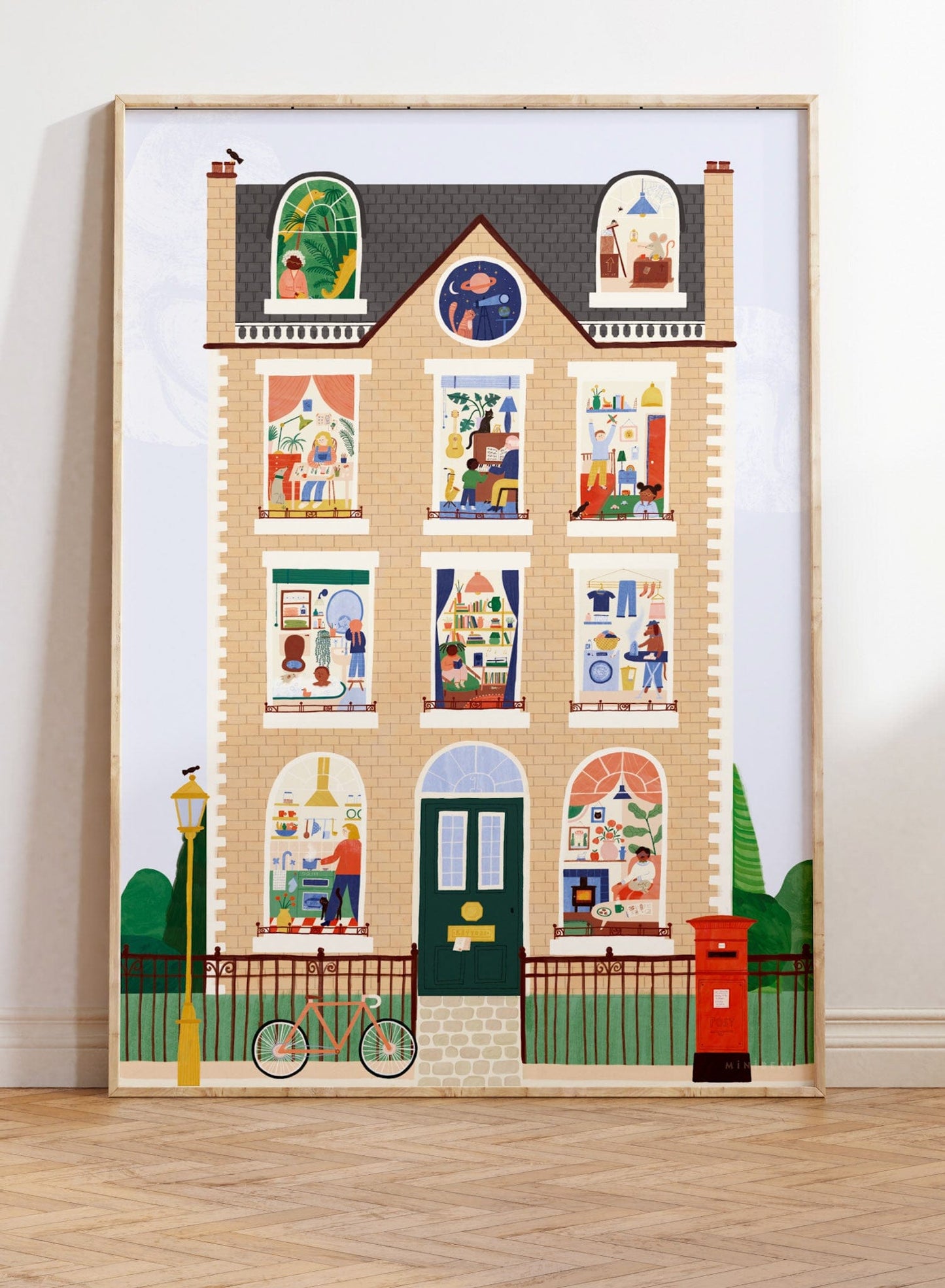 A large art print leaning against a white wall, stood on parquet flooring. Our House Art Print shows a large brick town house with 11 windows each showing a different scene, from an ironing dog to a dinosaur in the attic to the artist herself hard at work. Features a bike against a railing, a post box, traditional lamppost and hidden birds.