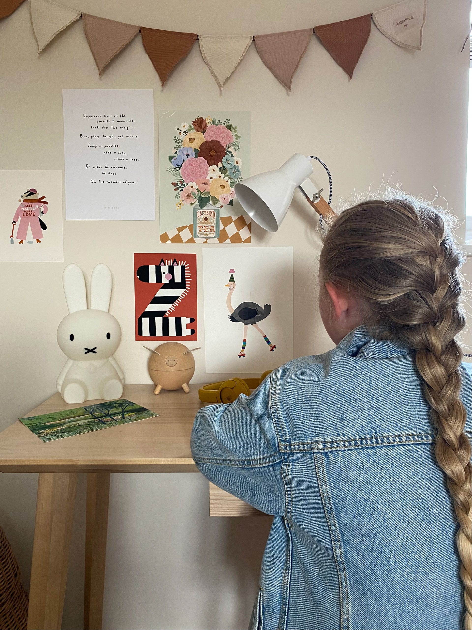 Girl sitting at her desk with a Miffy lamp on it with a selection of art prints stuck to the wall under Nobodinoz rose bunting. Featuring our boho floral vase art print, less plastic more love art print, Happy alphabet 'z' art print, party ostrich art print on the wall and the Garden art print on the desk.