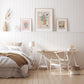 A scandi style bedroom showing a comfy bed with a white barrel night stand, and a white and light wood trestle table desk with a white chair and basket on the floor. On the shelf behind the bed are 3 art prints in light wood frames, the large print in the centre being our wild, wonderful, world leopard art print, and either side are smaller art prints, our oh, the places you go and mercado de flores.