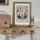 Showing one side of a wooden shelf with hanging pegs underneath with a wrapped wire word sign - HELLO, hanging on them. On the shelf is a Wooden flag saying Autumn, a wooden mouse, and Olli Ella Casa Clutch and our Mercado de flores art print in an oak frame.