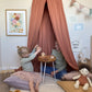 Two girls sitting on floor cushions under a rust coloured canopy, having a tea party with a wooden tea set from liewood and an Olli Ella doll in the foreground with the Boho Floral Vase art print, the wonder of you art print, and my wish for you art print on the wall behind.