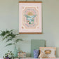 The top of a vintage iron fire place, with a white mantle, with boho style accessories on , including a miniature suitcase and a book. Hanging from a picture hook in an oak hanger is our oh the places you'll go art print. giving you are my sunshine vibes. 