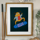 Our rad dog art print in an oak frame with a white mount low on a wall in a kids play area. with blue raindrop bunting, a vintage wooden toy race car on a wooden train track and a Liewood Mary wooden gold set. Our Rad Dog Art Print features a brown dog doing a flip on a skateboard. The dog is wearing a denim blue jacket and a bright pink and orange top while wearing black and white striped glasses on a dark green background.