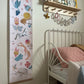 Our folk carnival height chart hanging on a neutral wall. Vintage metal framed bed in the background and a wooden shelf with peg hooks with a miffy lamp on it and raindrop bunting.