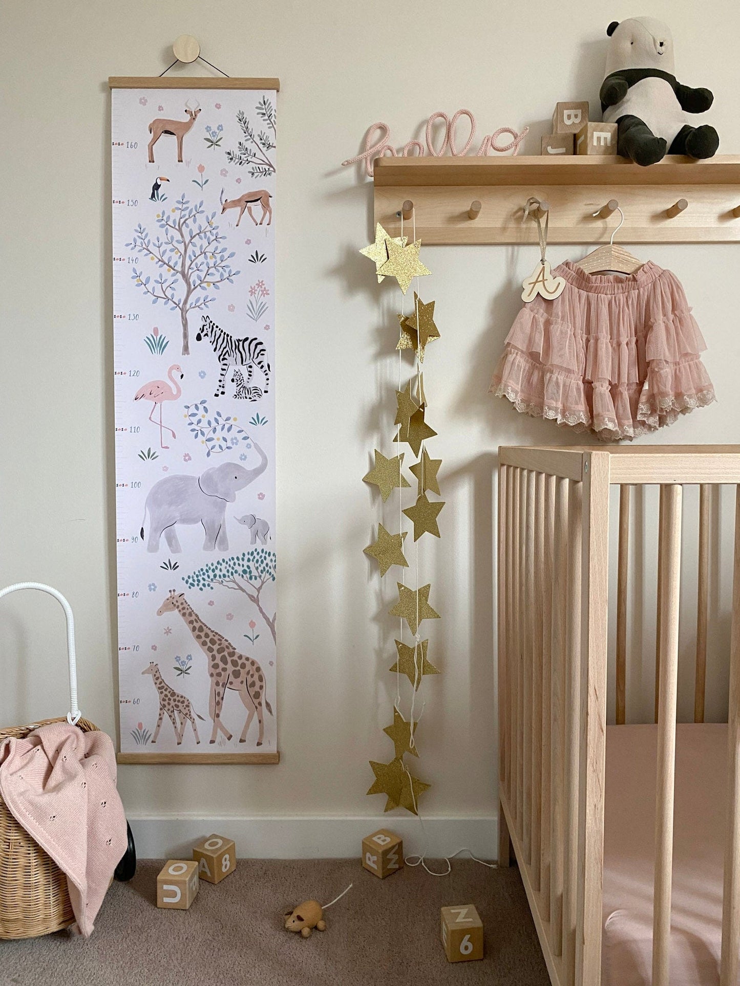 Our Serengeti height chart with oak hanger hung from a round wooden hook on a wall next to a cot with a star garland hanging down from a shelf with hooks underneath. On the shelf is a hello kiddo hello sign, a plush panda and some blocks